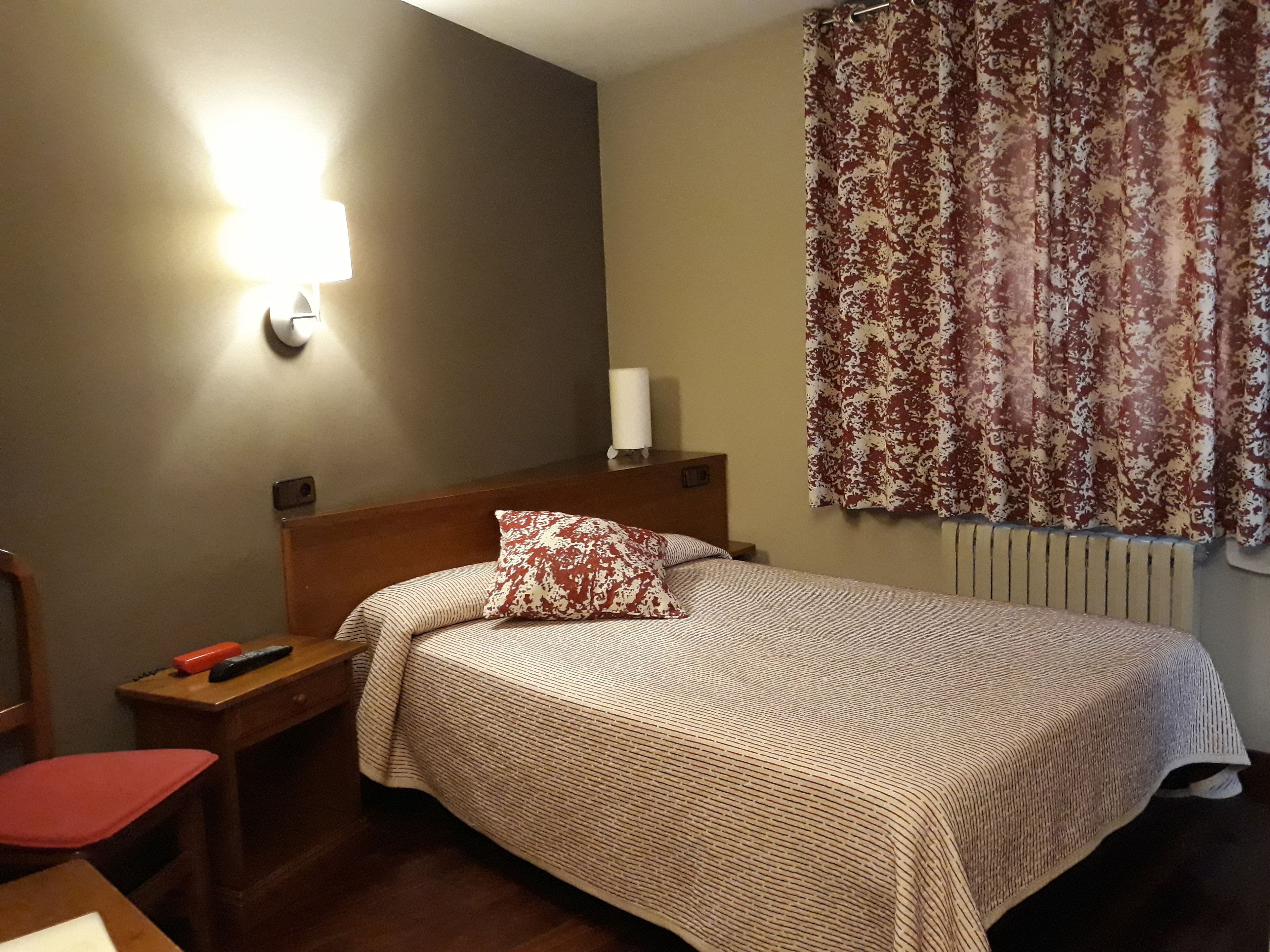 A hotel room with a bed, a nightstand, a lamp, a chair, and a window with curtains.