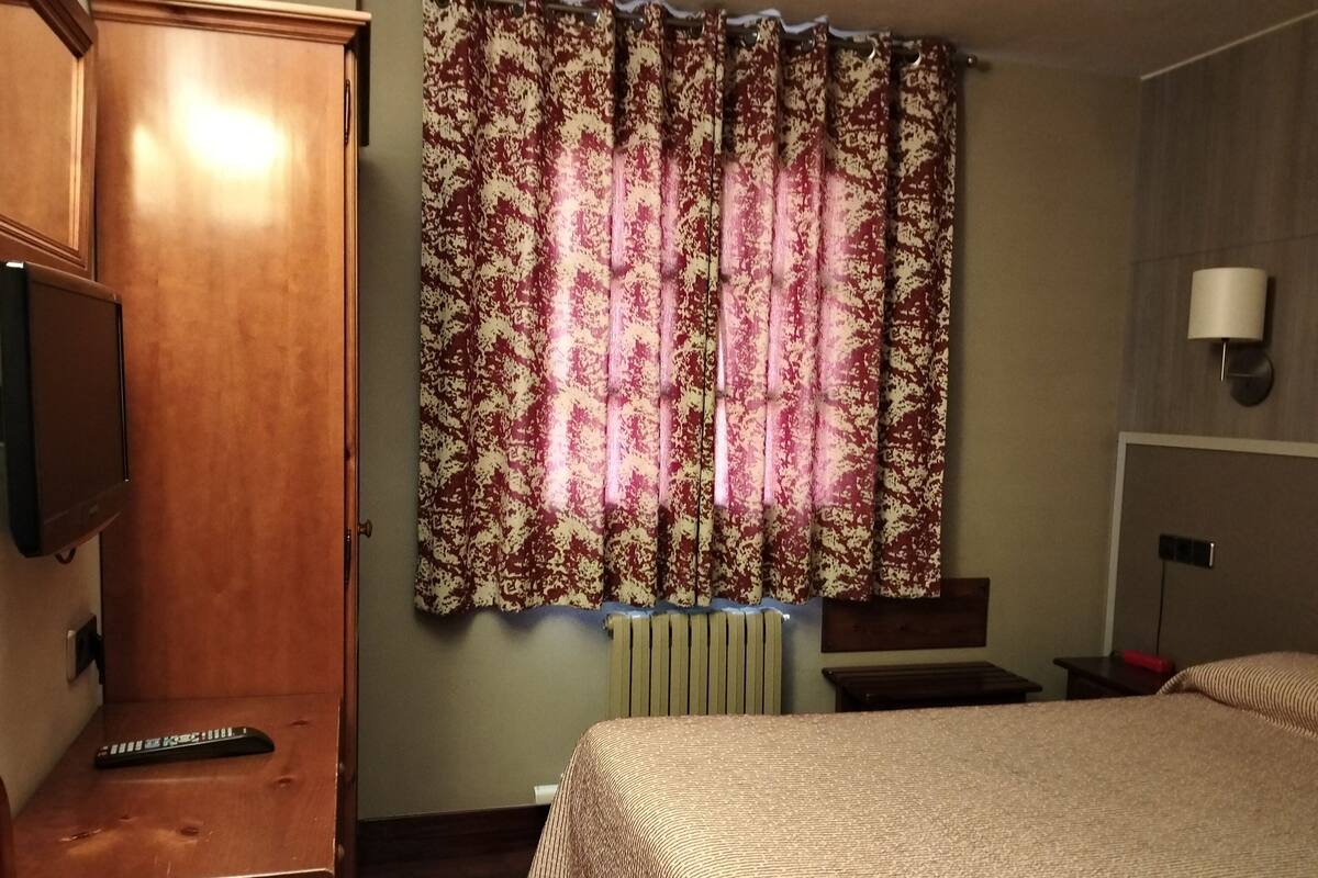 Una habitación de hotel con cama, un armario de madera, televisor y ventana con cortinas estampadas.
