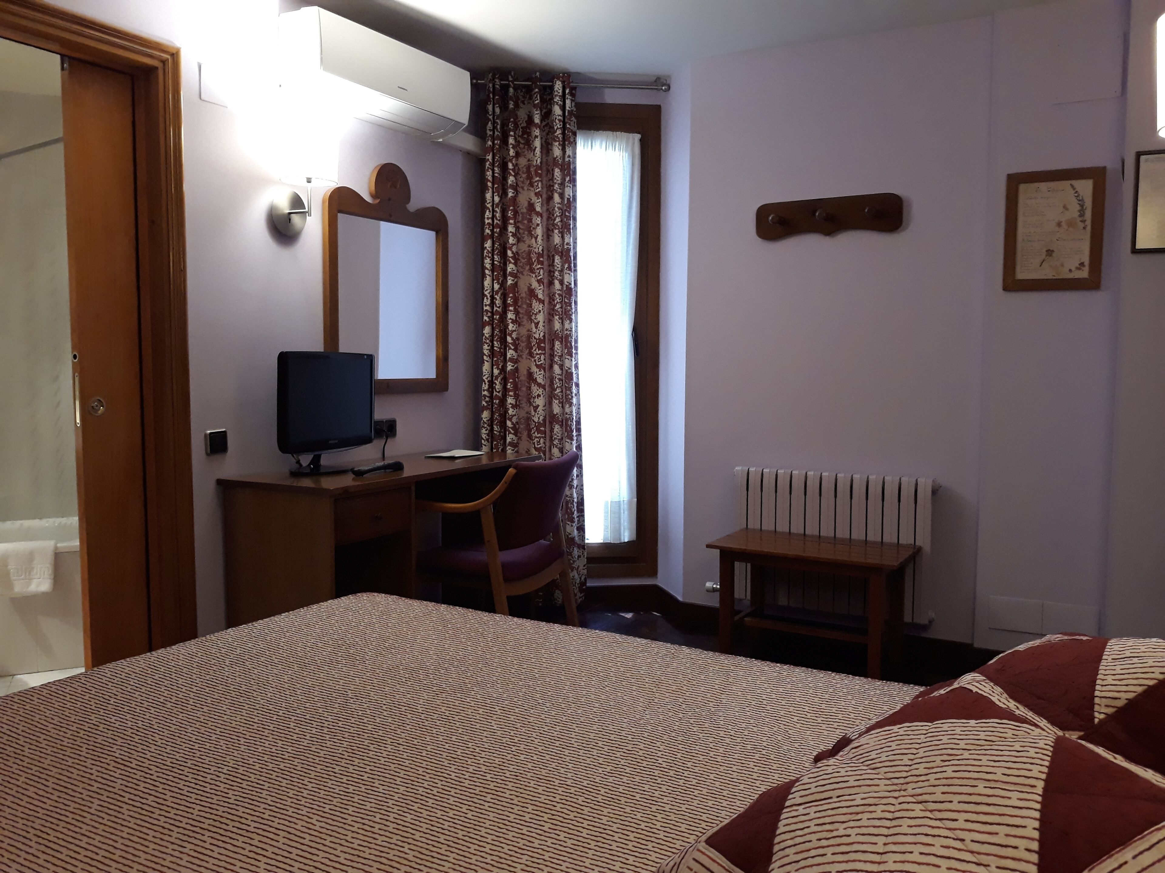 A hotel room with a bed, a desk with a computer, a chair, a radiator, and a window with curtains.