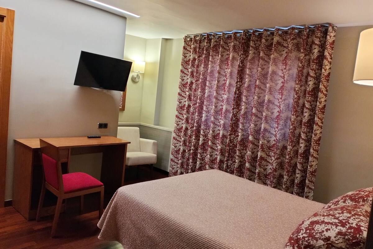 Habitación de hotel con cama, escritorio con silla, televisor montado en la pared y cortina con estampado floral.