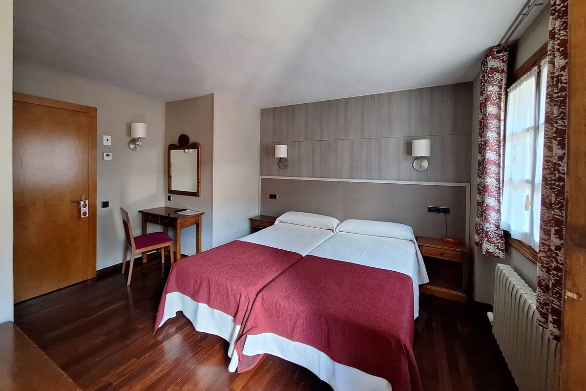 Habitación de hotel con piso de madera, cama con cubrecama rojo y blanco, escritorio con silla, espejo, dos lámparas fijadas a la pared y una ventana con cortinas.