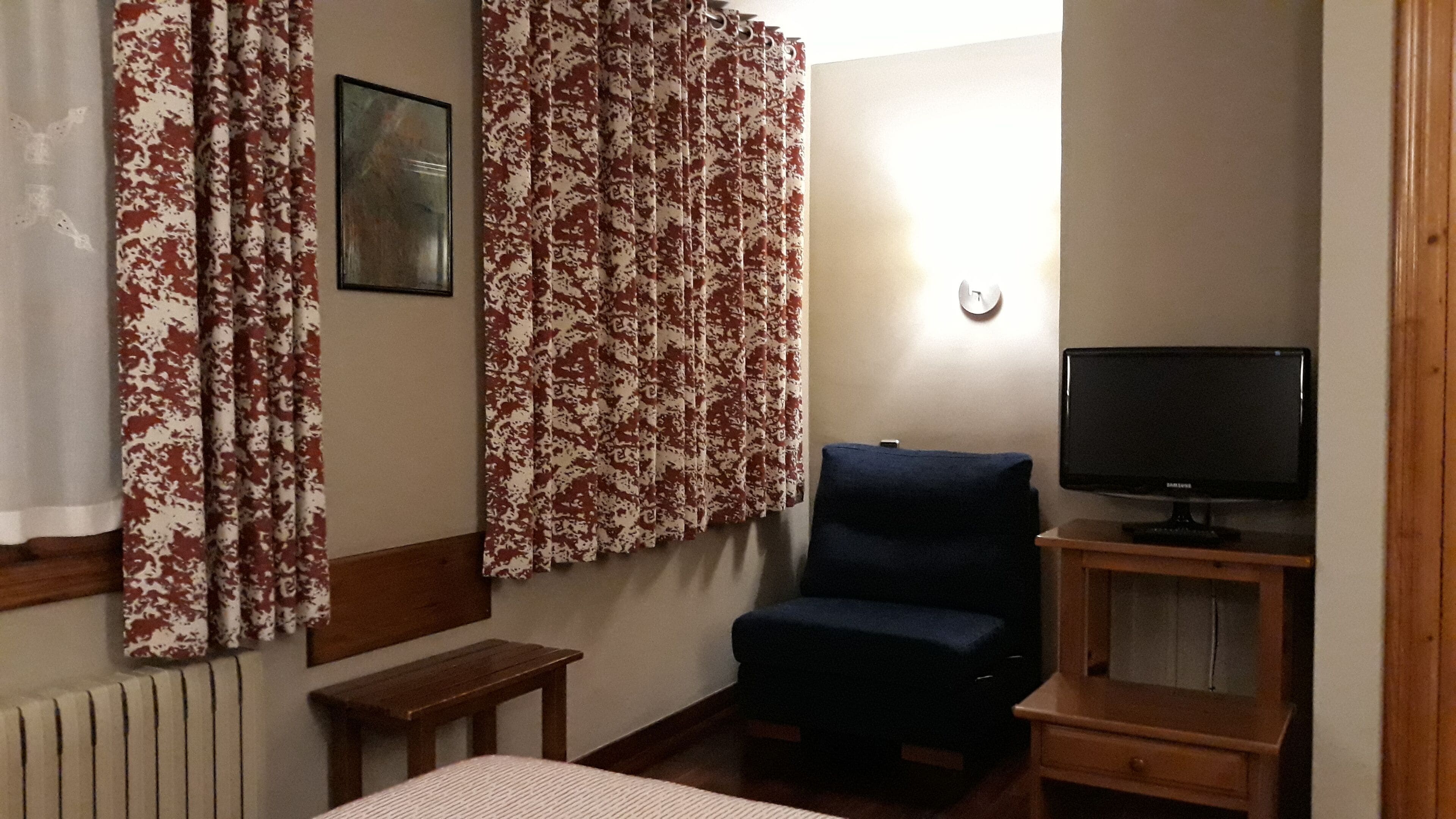 A room with a bed, a chair, a TV on a wooden stand, and patterned curtains.