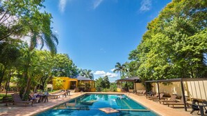 A swimming pool surrounded by lounge chairs and shaded areas, with trees and clear skies in the background. - Pirayu Lodge Resort (Puerto Iguazú)