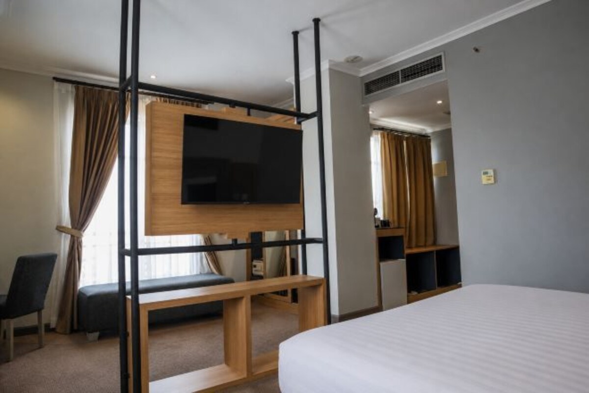 A hotel room with a bed, a TV mounted on a wooden stand, a desk, and a chair.