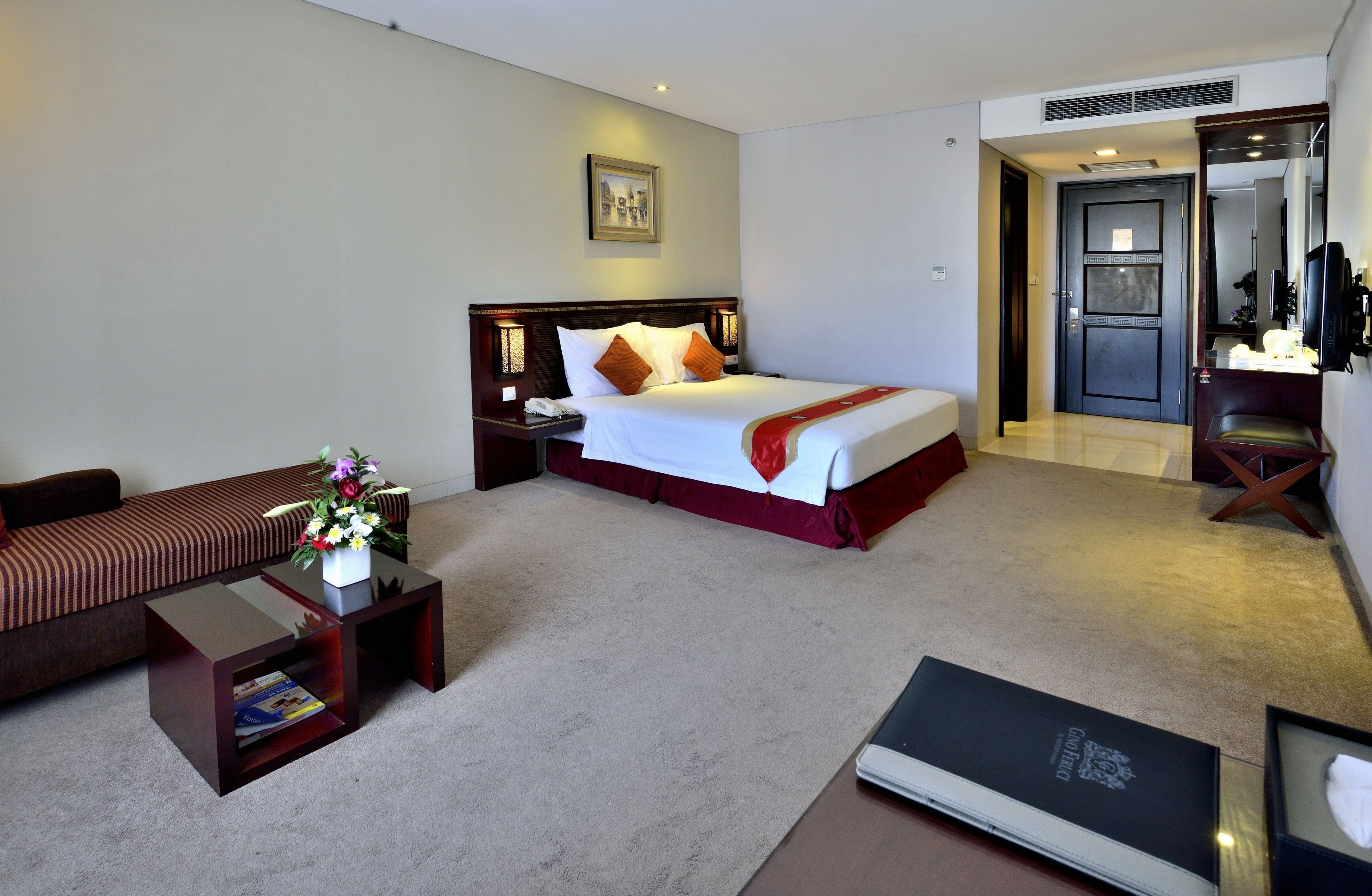 A hotel room with a bed, a sofa, a nightstand, a small table with a vase of flowers, and a door leading to a bathroom.