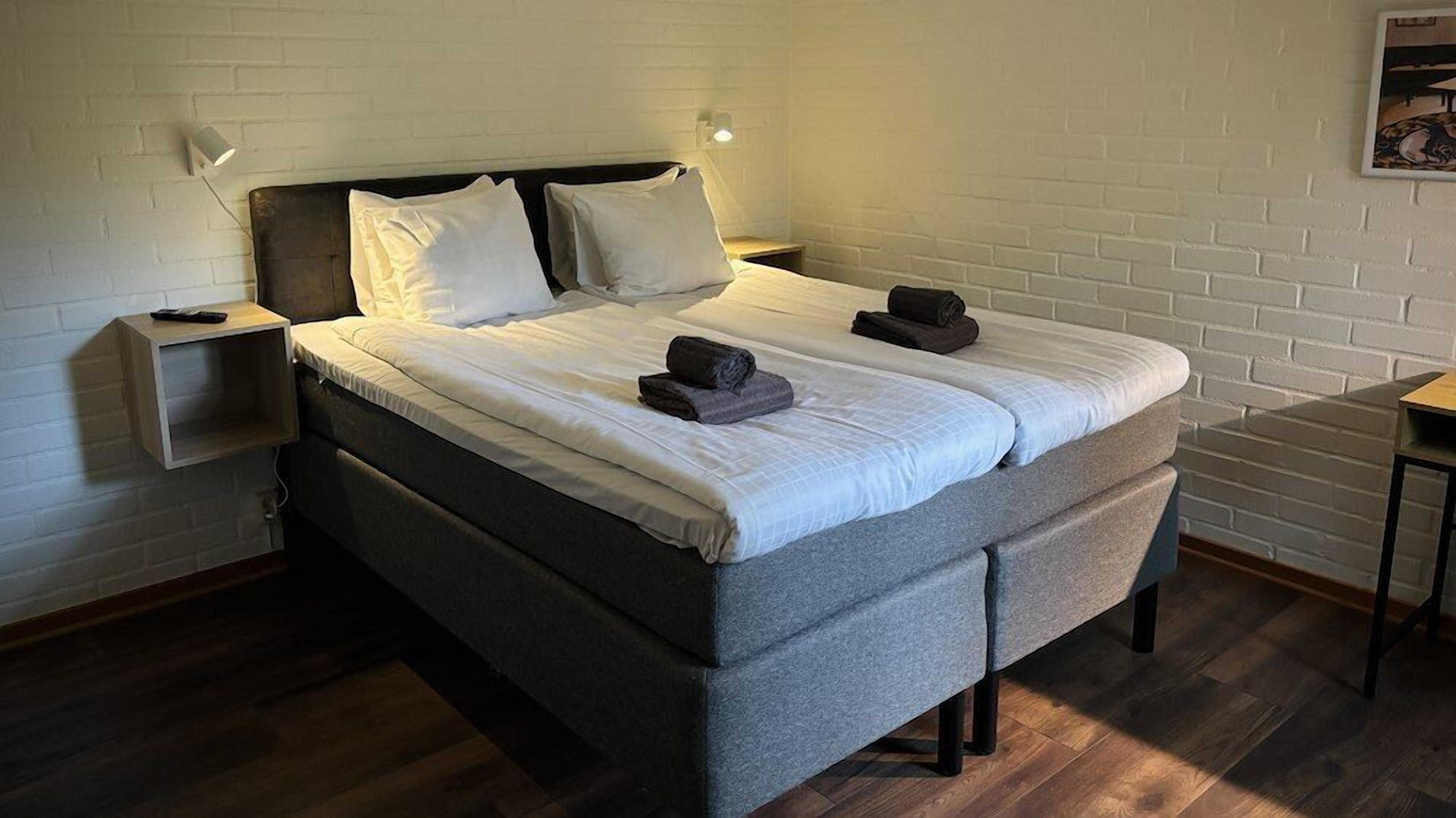 Ein Bett mit grauem Kopfteil, weißer Bettwäsche und zwei aufgerollten Handtüchern. Ein Nachttisch mit Lampe sowie ein kleiner Tisch mit Stuhl befinden sich rechts.