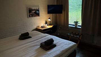 A hotel room with two beds, a desk with a lamp, a TV, and a window with a plant.