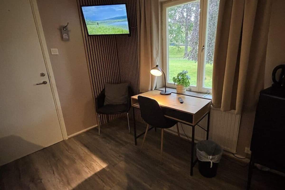 Ein kleines Zimmer mit Schreibtisch, zwei Stühlen, einem an der Wand befestigten Fernseher, einem Fenster mit Vorhängen und einer Heizung.
