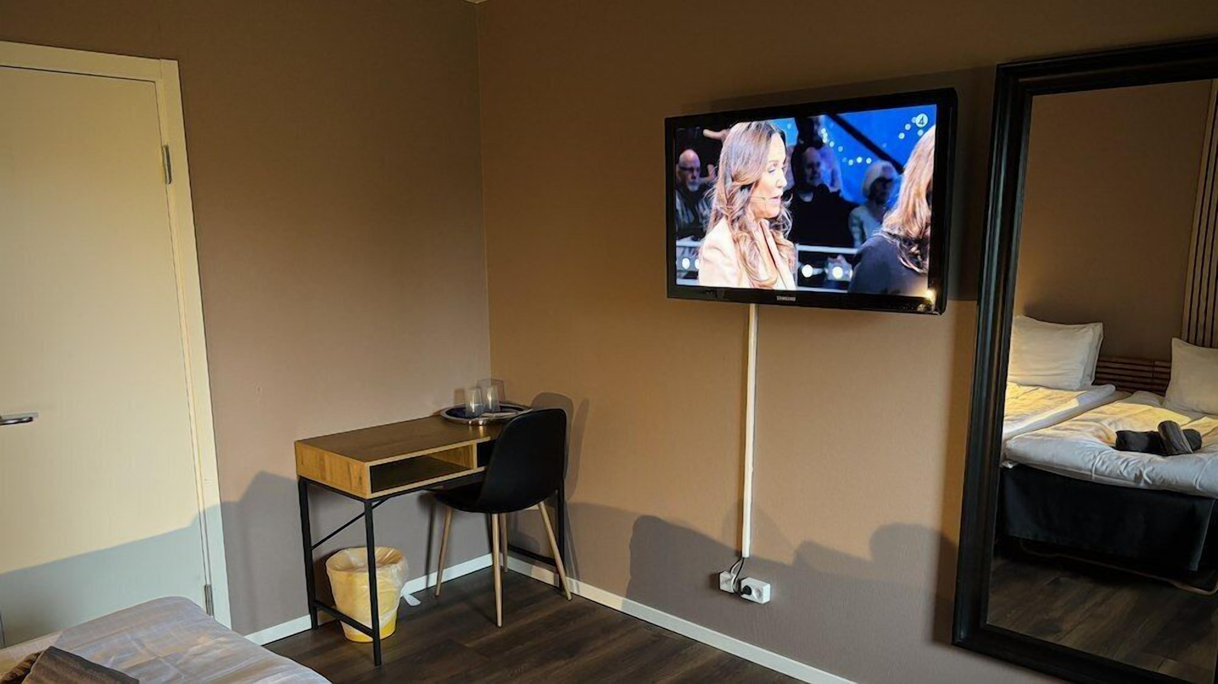 Habitación de hotel con cama, escritorio con silla, televisión montada en la pared y un cesto de basura.