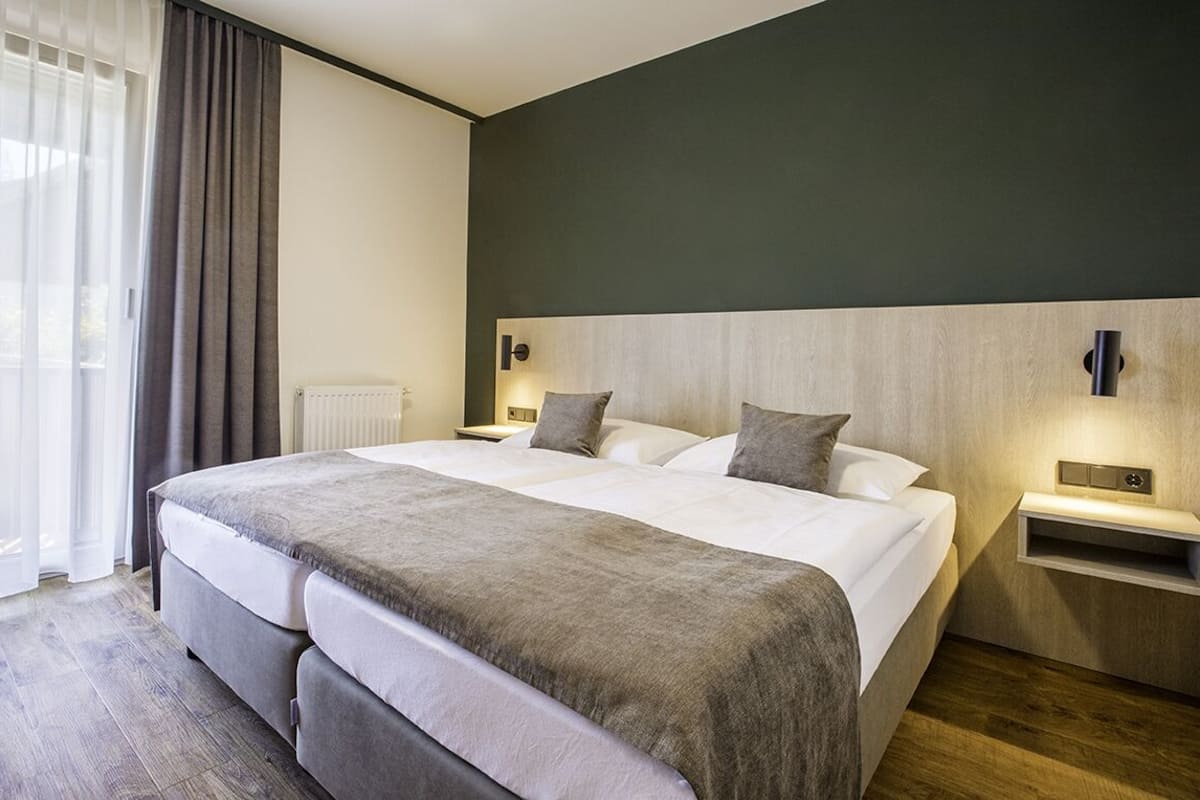 A modern hotel room with a large bed, a bedside table, a chair, and a view of the outdoors through sheer curtains.
