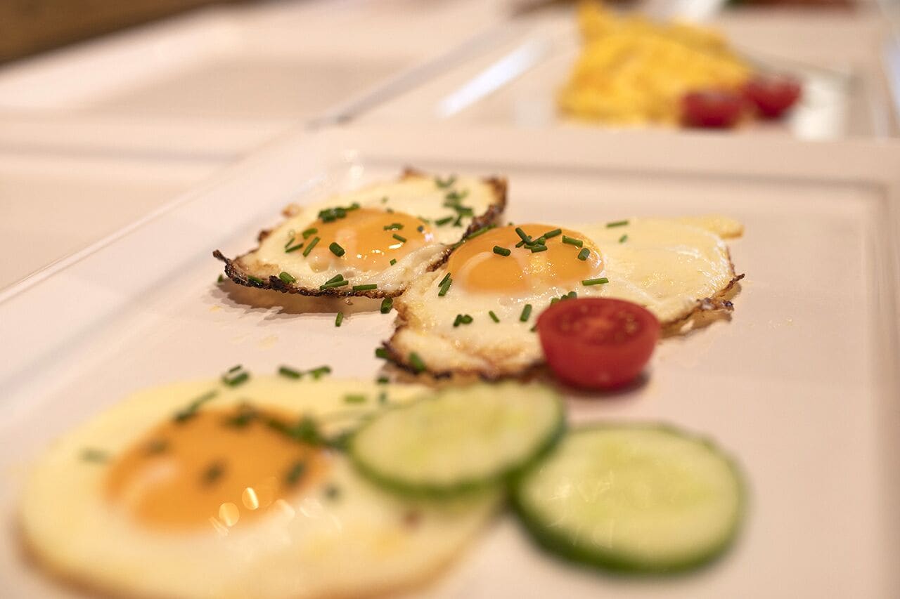 A plate with three poached eggs on toast garnished with chives, accompanied by slices of cucumber and a cherry tomato.