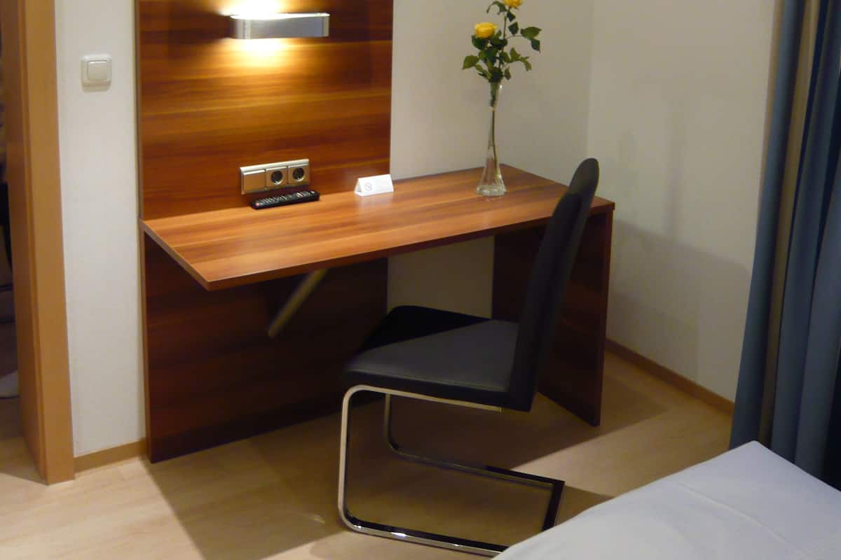 A hotel room with a wooden desk, a television mounted on the wall, a vase with flowers, a chair, and a bed.