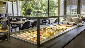 A buffet area with various food options, including fruits, cheeses, and desserts, in a modern restaurant setting with large windows and natural light.