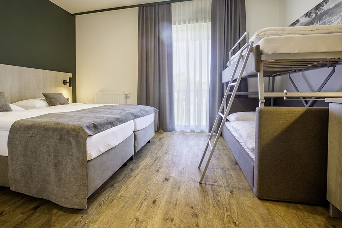 A hotel room with two beds, a bunk bed, wooden flooring, a radiator, and a window with curtains.