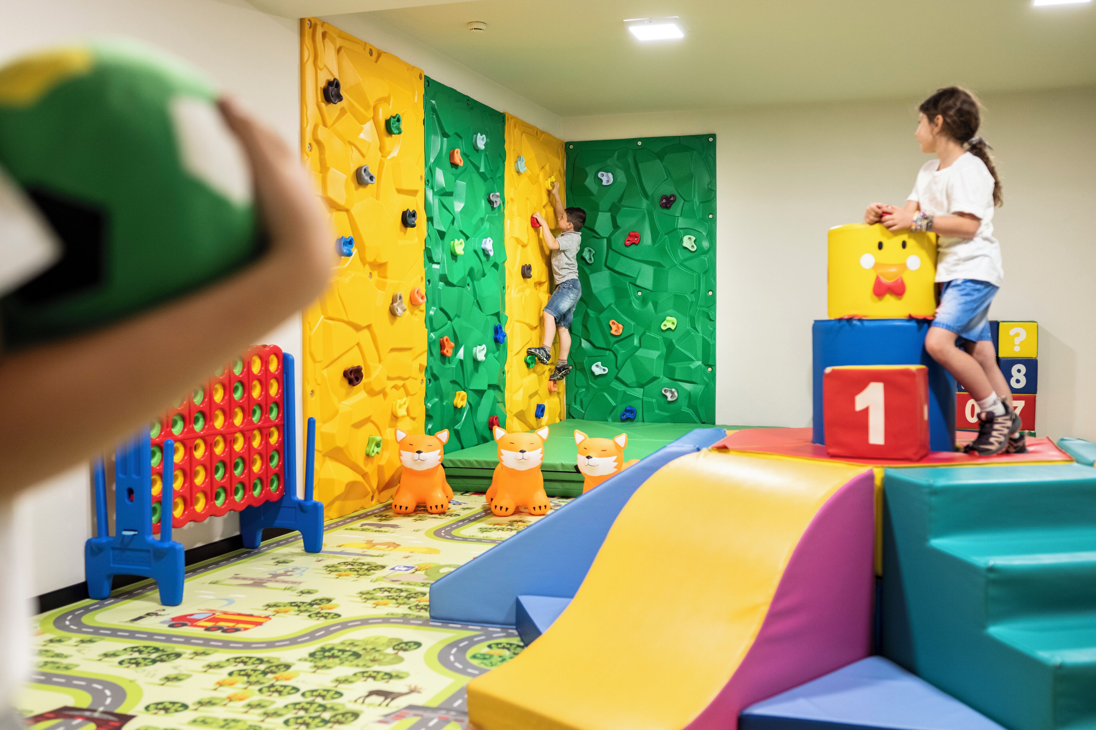 A child climbing a colorful indoor climbing wall, a soft play area with a slide, and a play mat with cartoon characters.