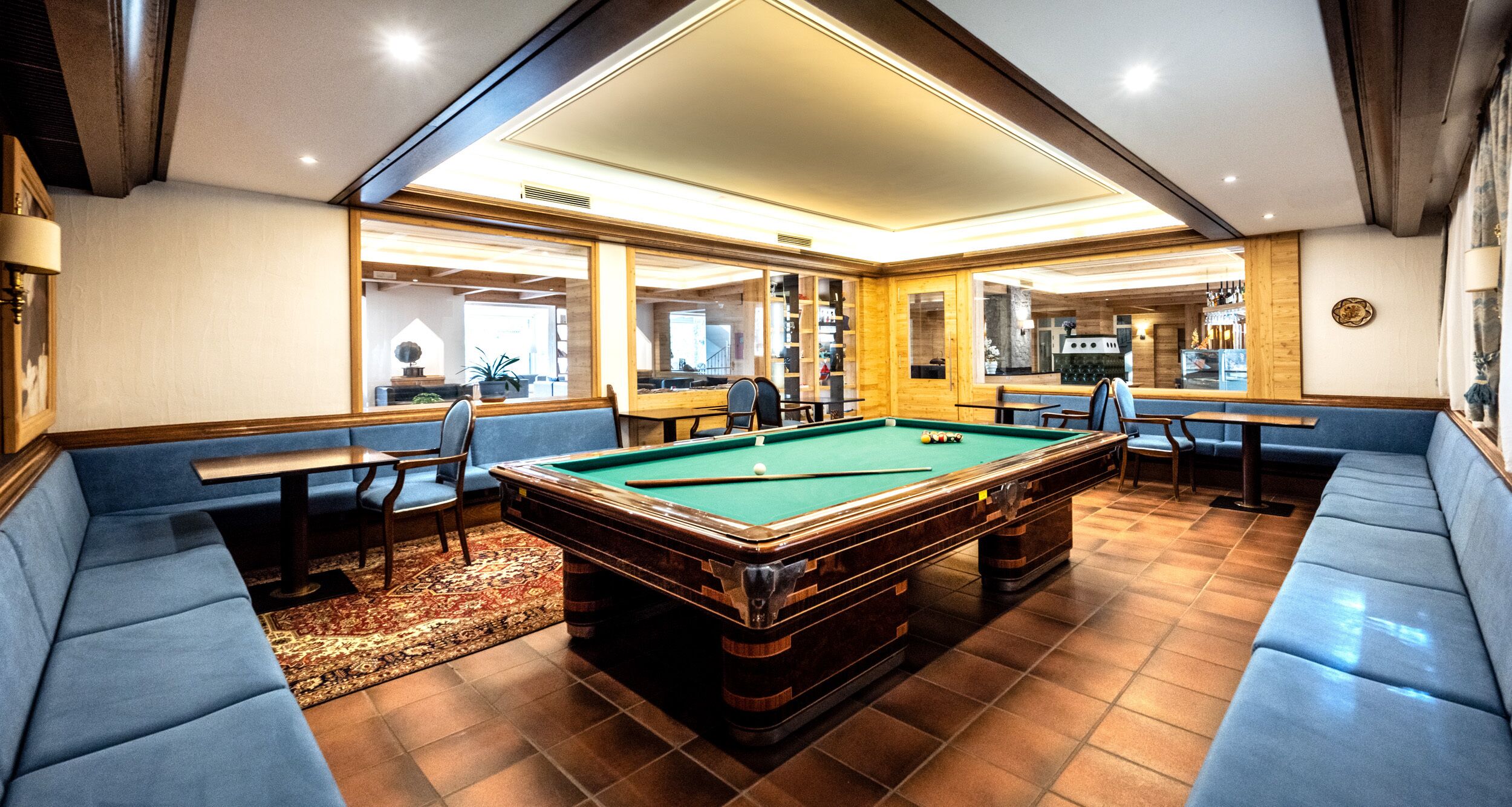A pool table in a room with blue seating and a kitchen area visible through glass doors.