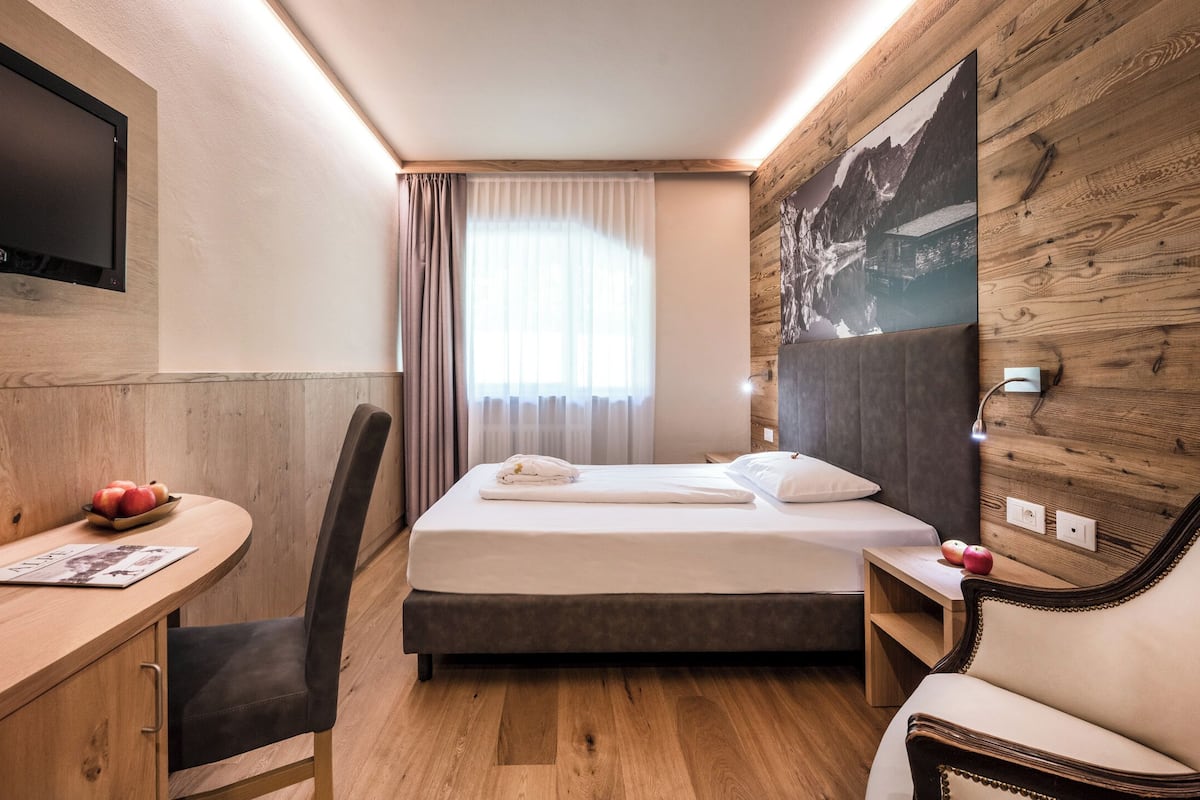 A hotel room with a bed, a desk with a chair, a TV mounted on the wall, and a large window with curtains.