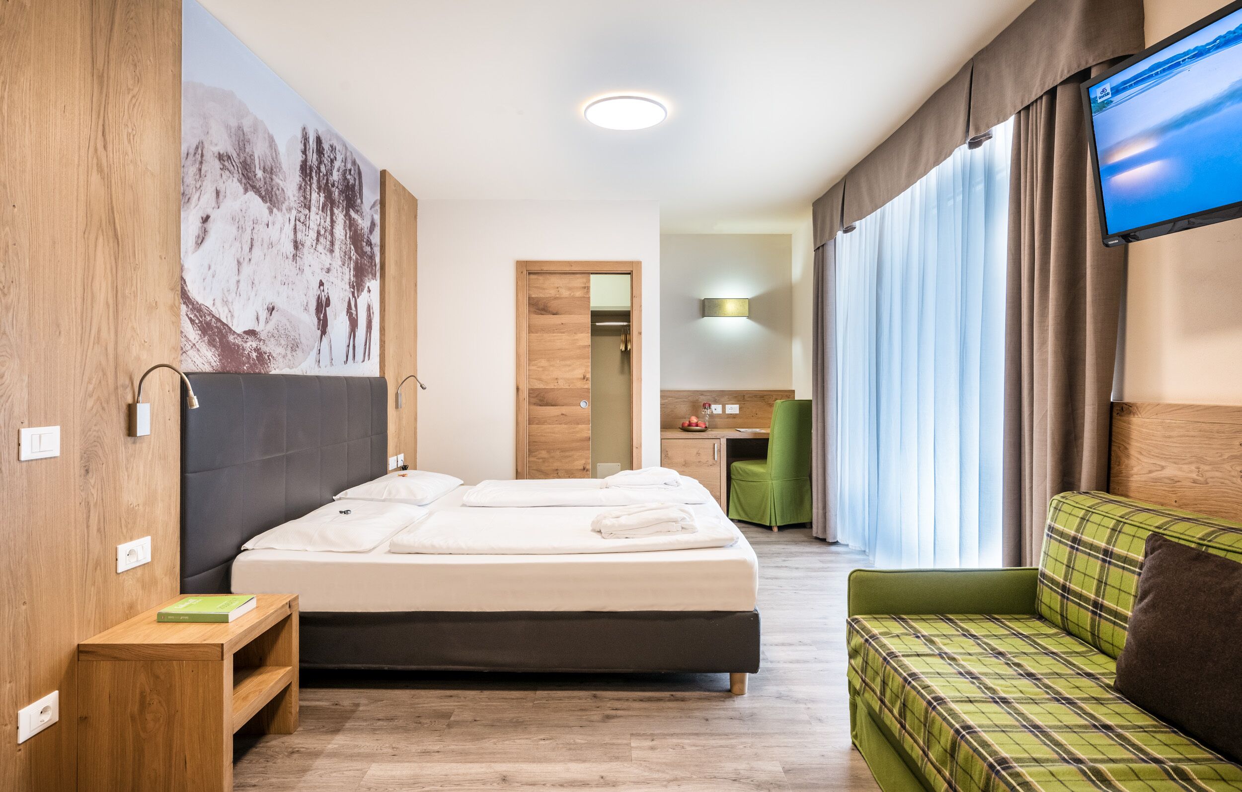 A modern hotel room with a large bed, a green sofa, a flat-screen TV, and a wooden accent wall.