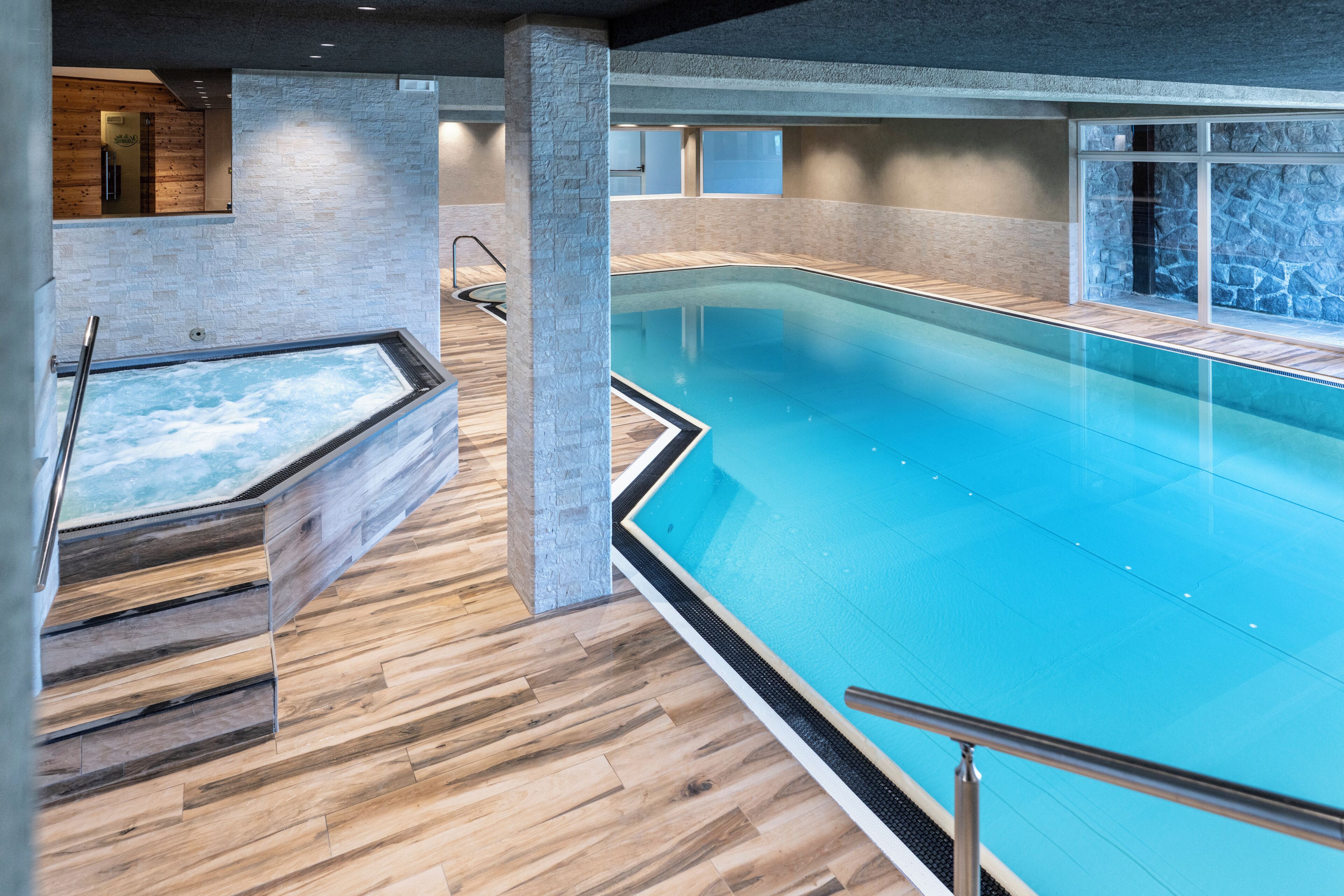 A large indoor swimming pool with a wooden deck area, stone walls, and a view of the sky.
