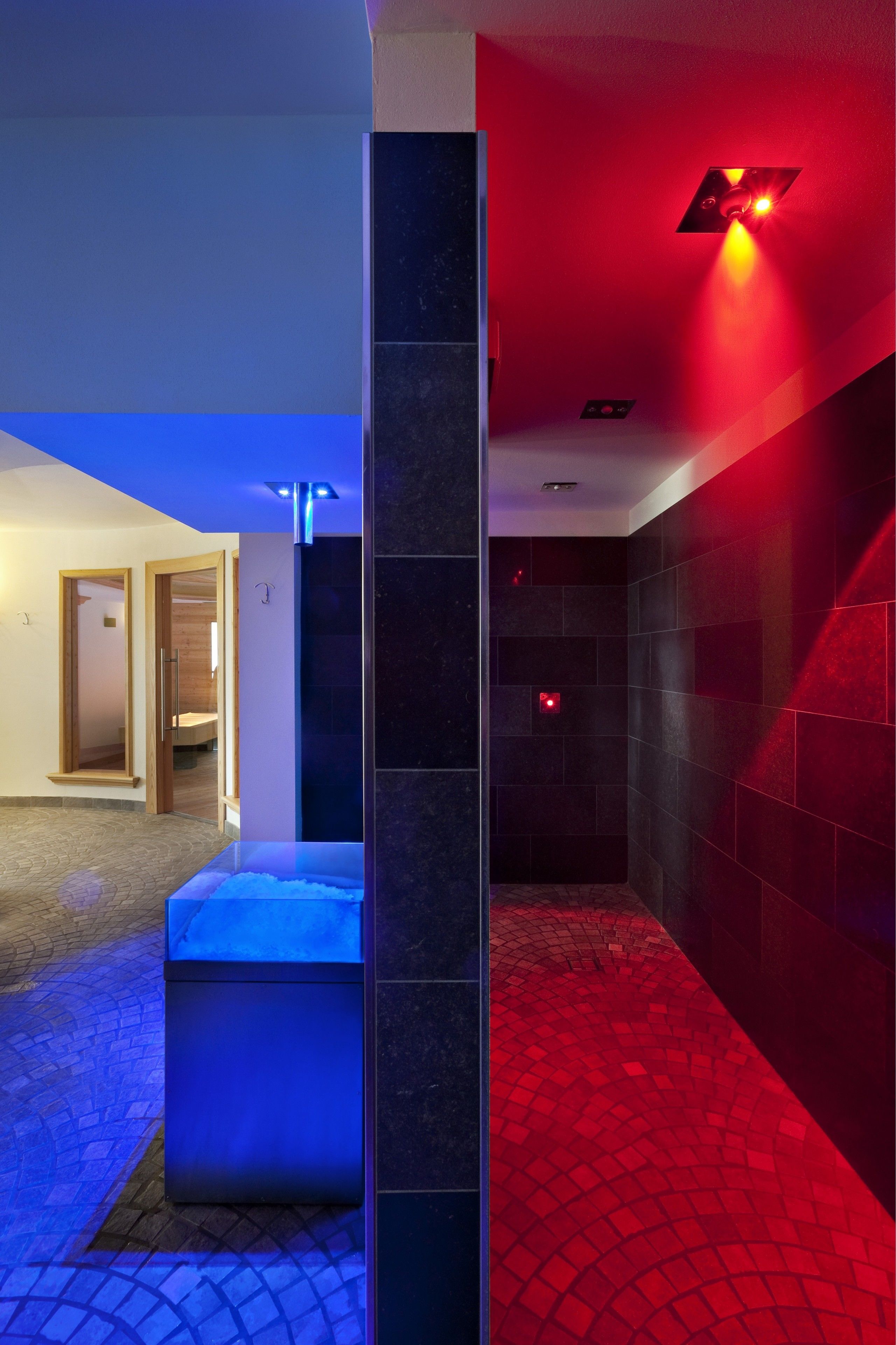 Ein modernes Badezimmer mit einer blau beleuchteten Waschtisch-Konsole, einem rot beleuchteten Duschbereich und einer sichtbaren Tür zu einem weiteren Raum.