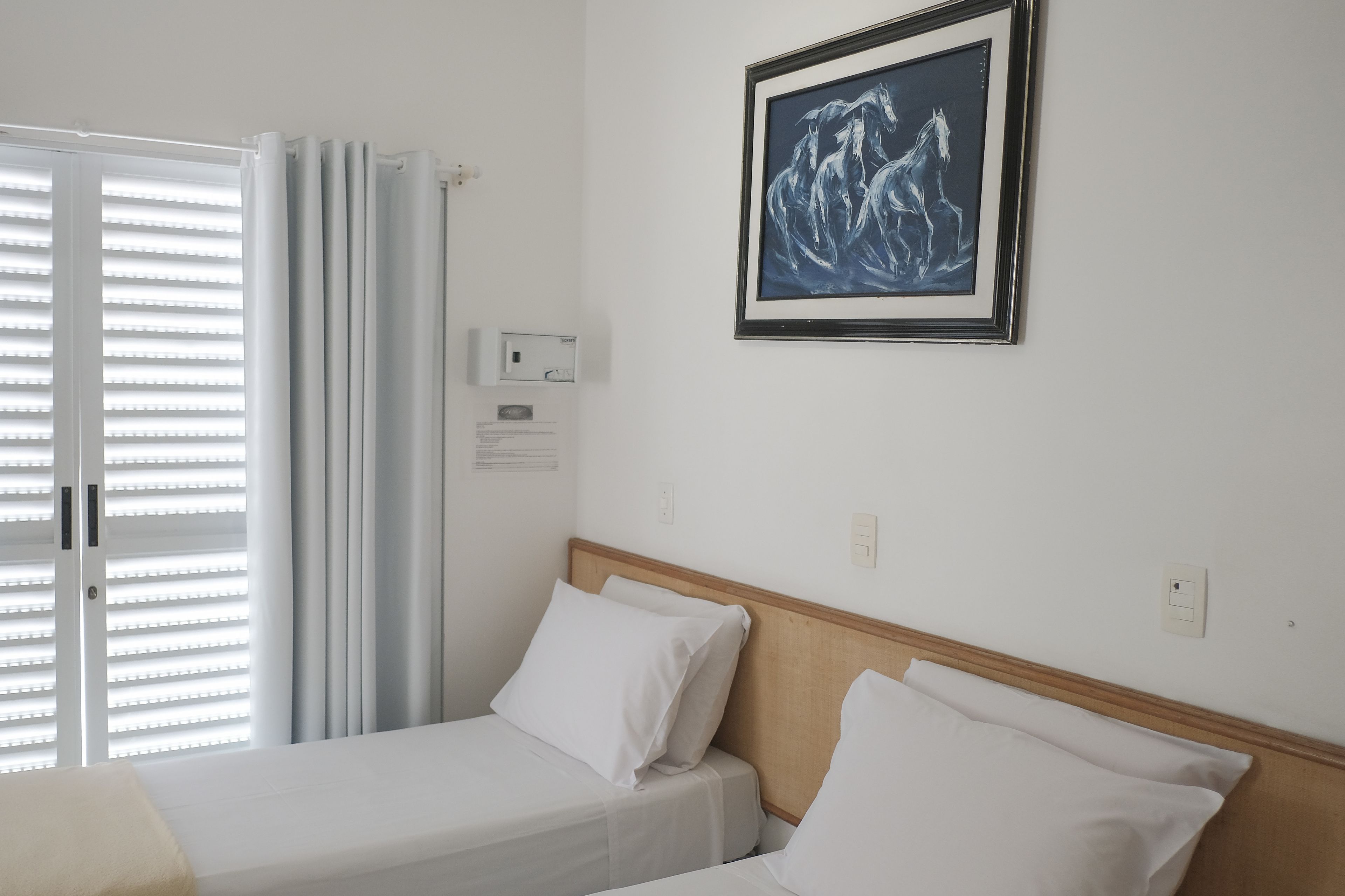 A hotel room with a bed, white bedding, a framed artwork on the wall, and a window with blinds.