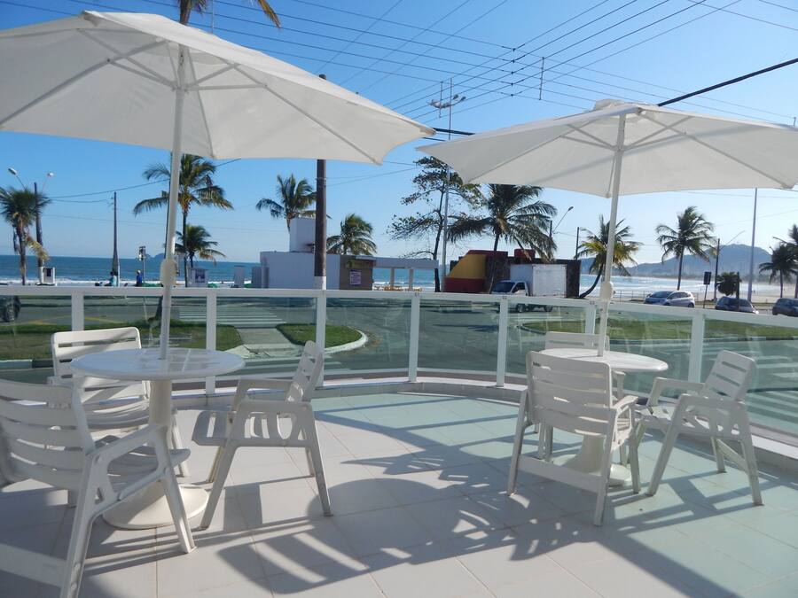 Terraço com cadeiras brancas e guarda-sóis, com vista para uma praia e palmeiras.