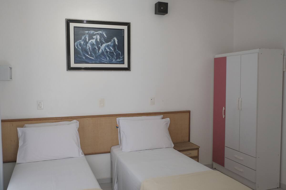 A hotel room with two single beds, a wooden headboard, a white wardrobe, and a framed artwork on the wall.