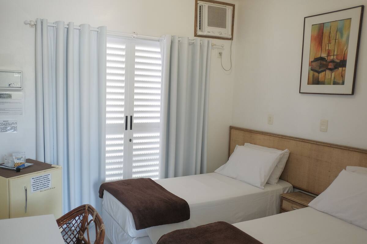 A hotel room with two beds, a desk, a chair, a radiator, and a painting on the wall.