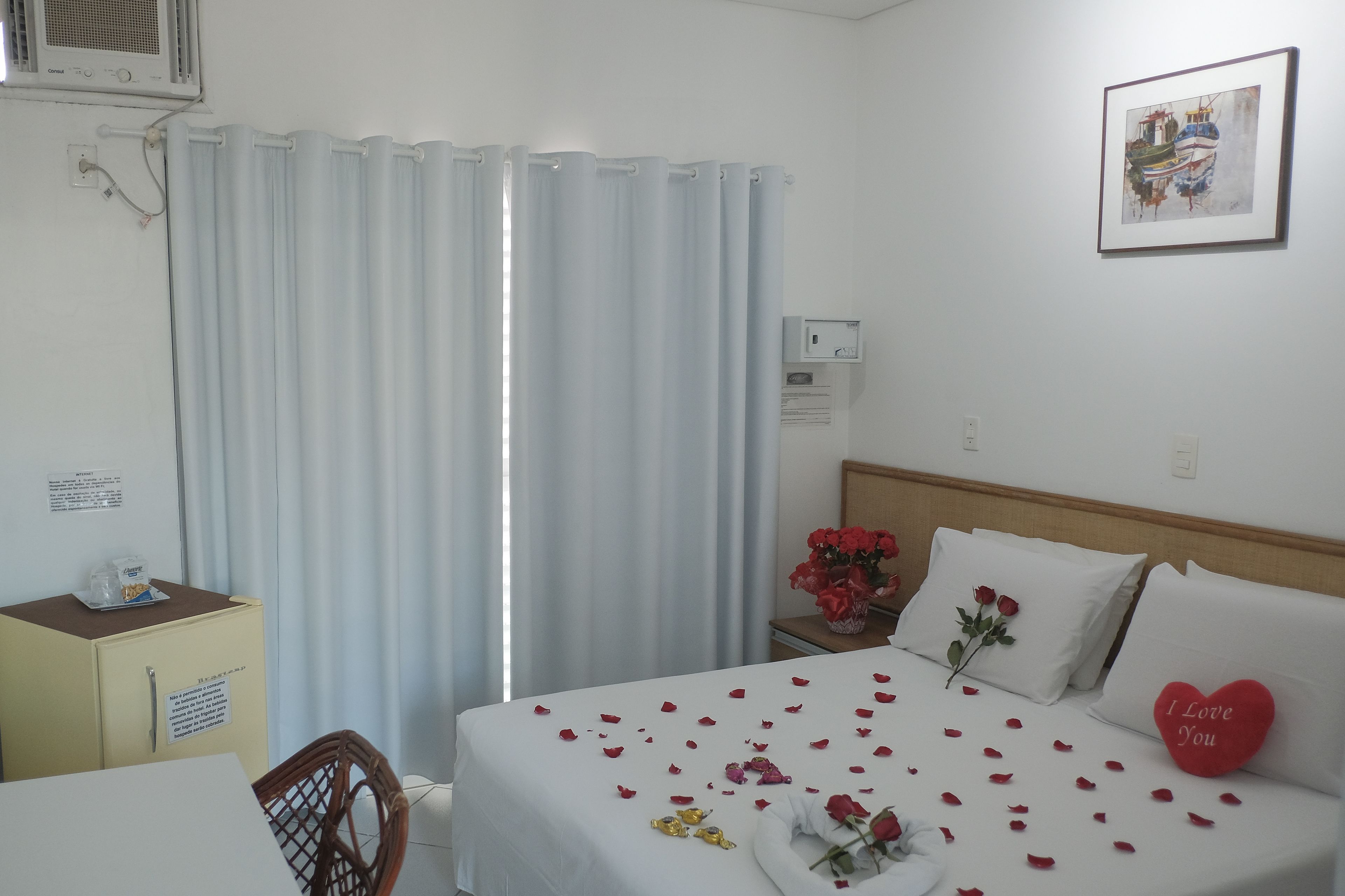 A hotel room with a bed, a bedside table with a vase of red flowers, a red heart-shaped pillow, and a framed picture on the wall.