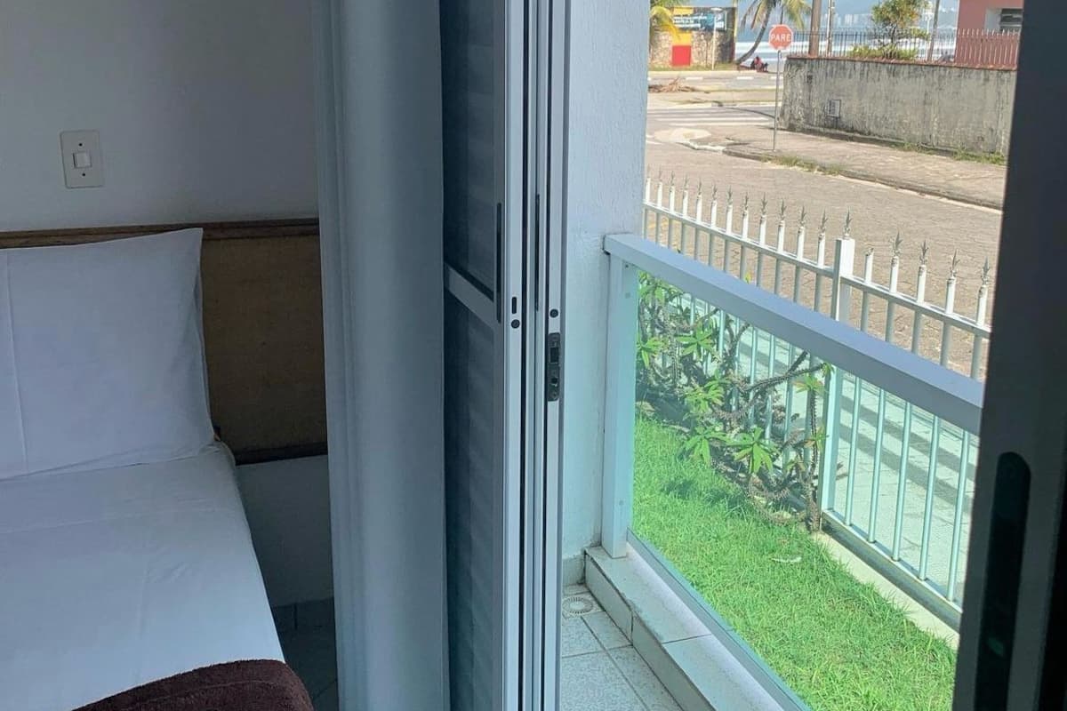A hotel room with a bed, a window with a view of a street and palm trees, and a balcony with a railing.