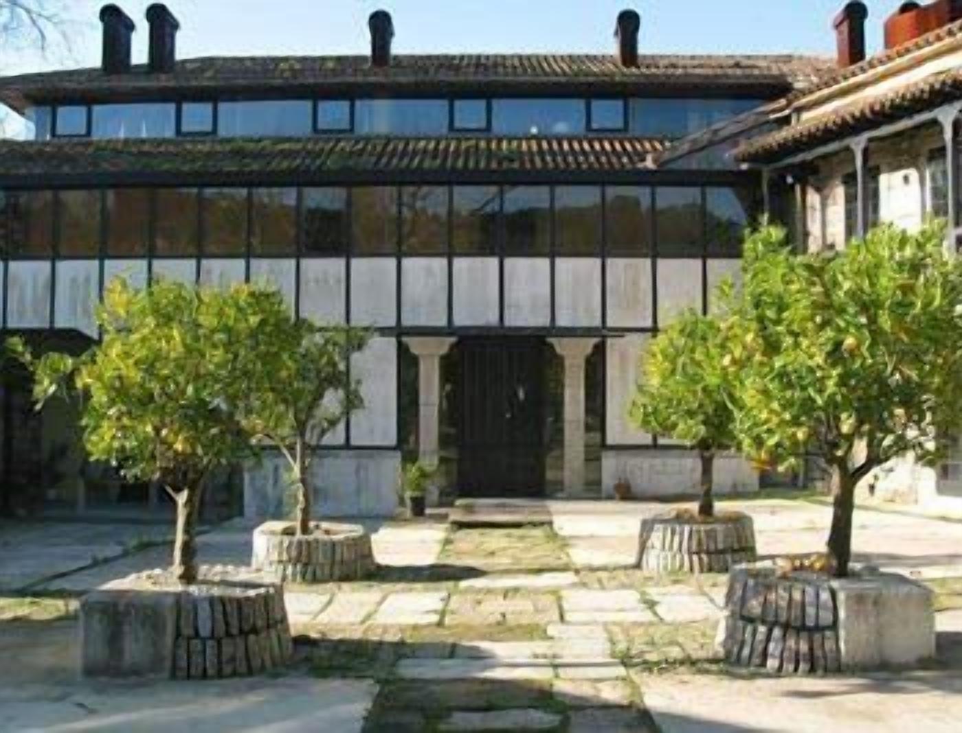 Un bâtiment à la façade symétrique, doté de grandes fenêtres et d’une entrée centrale flanquée de colonnes. Deux arbres sont plantés dans des jardinières devant l’entrée.