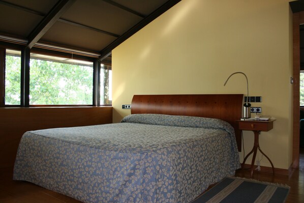 A bedroom with a large bed, a wooden headboard, a bedside table with a lamp, and a window with a view of greenery.