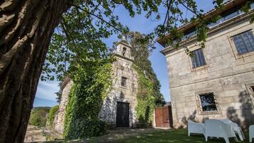 Un edificio de piedra con un campanario, muros cubiertos de hiedra y un patio césped con sillas blancas.