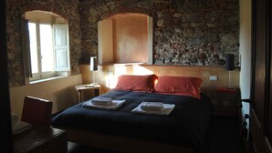 Un dormitorio con pared de piedra, una cama con almohadas rojas, una mesita de noche y una ventana con cortinas.