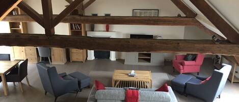 A spacious loft with exposed wooden beams, a central seating area with sofas and a coffee table, and a kitchen in the background.