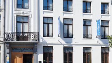 Un bâtiment blanc avec une enseigne indiquant « Royal Astrid Hotel ». Il possède de nombreuses fenêtres, un balcon et une zone d’entrée pavée agrémentée de plantes en pot.