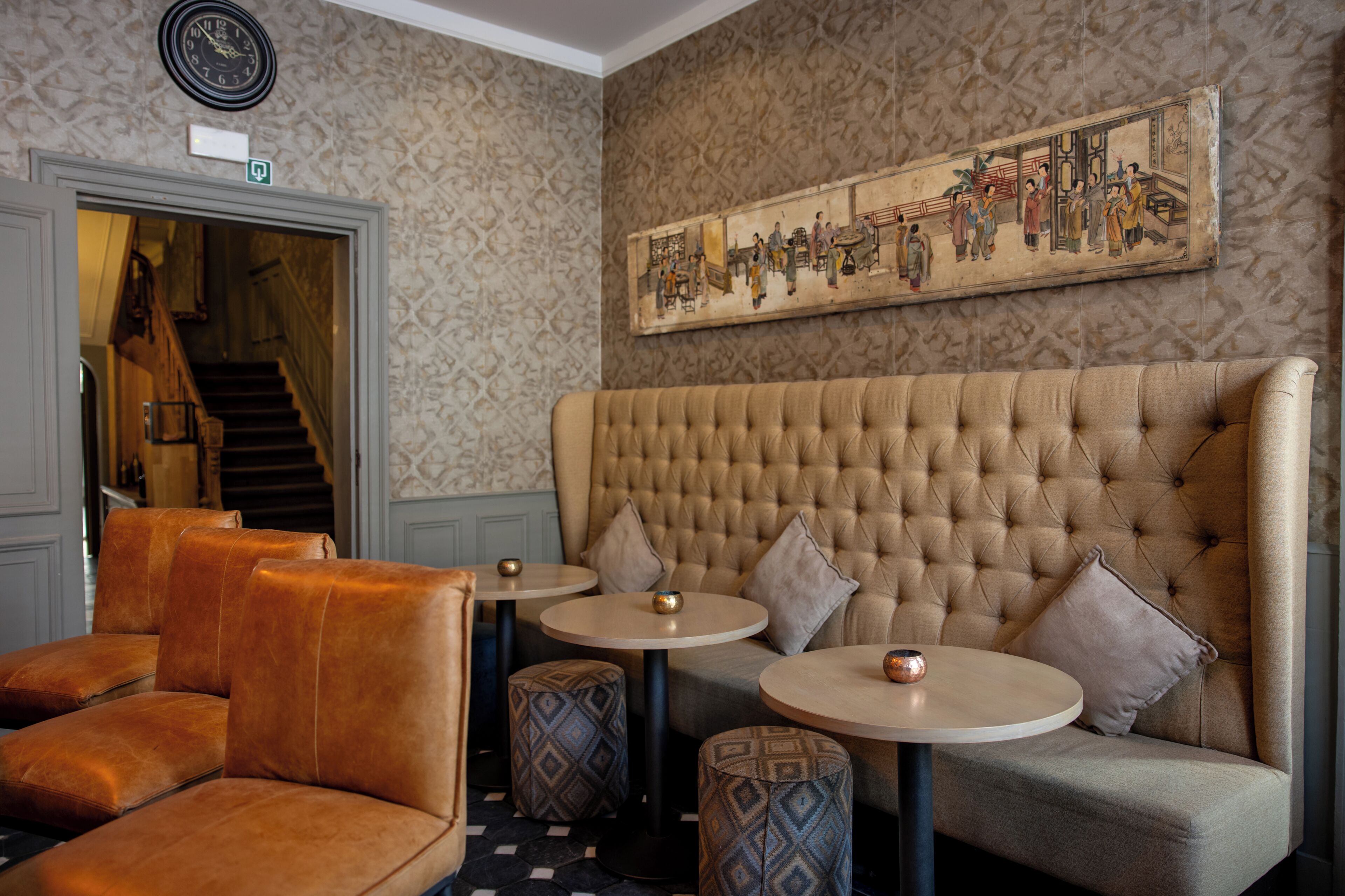 A lobby with a tufted sofa, round tables, patterned wallpaper, a clock, and a traditional painting.