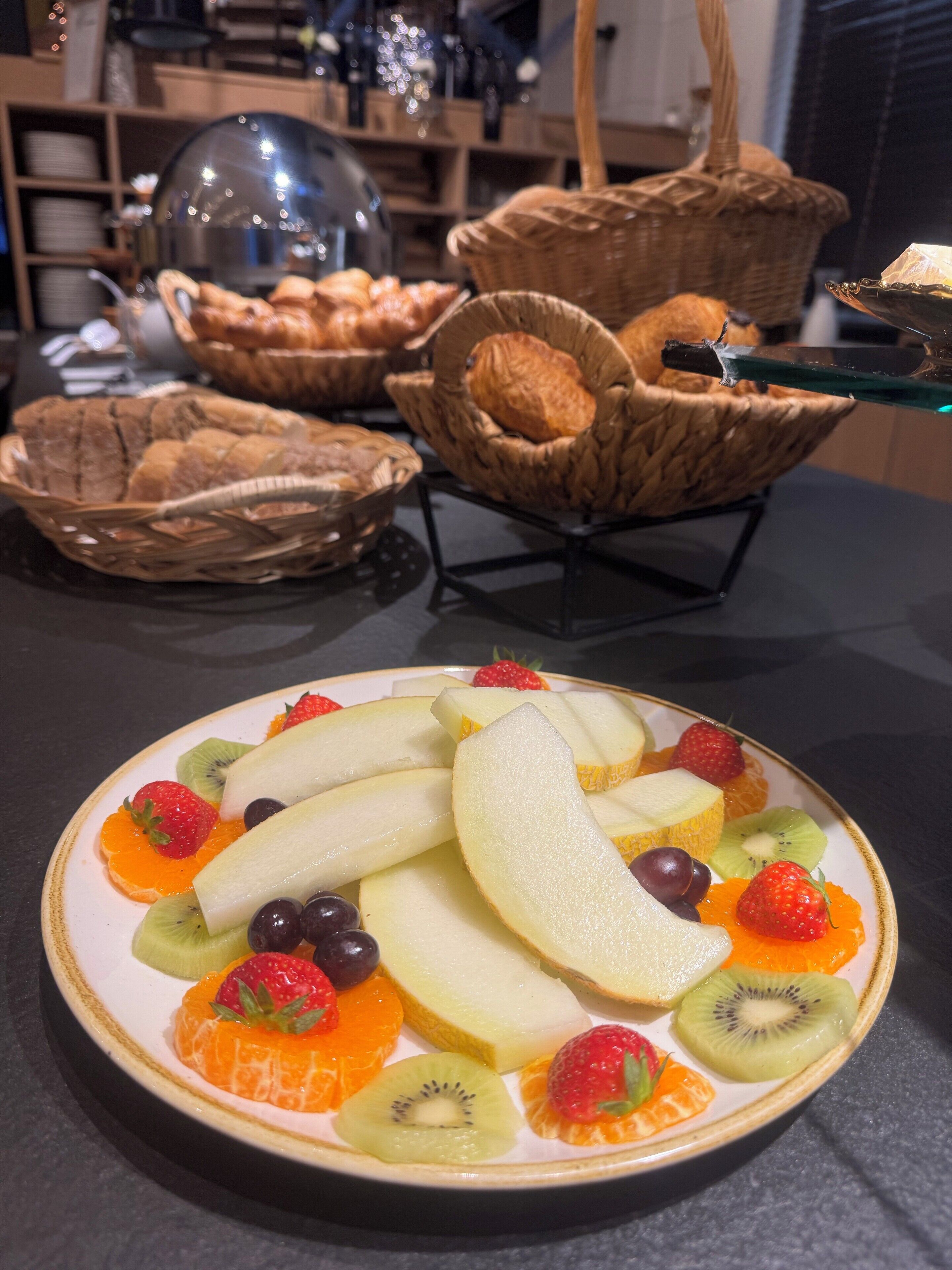 Uma travessa de frutas sobre a mesa, com cestas de pão ao fundo.