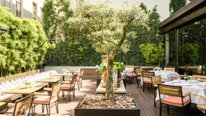 An outdoor dining area with wooden tables and chairs, surrounded by lush greenery and a central tree. - Square Nine Hotel Belgrade-The Leading Hotels of The World (Belgrade)