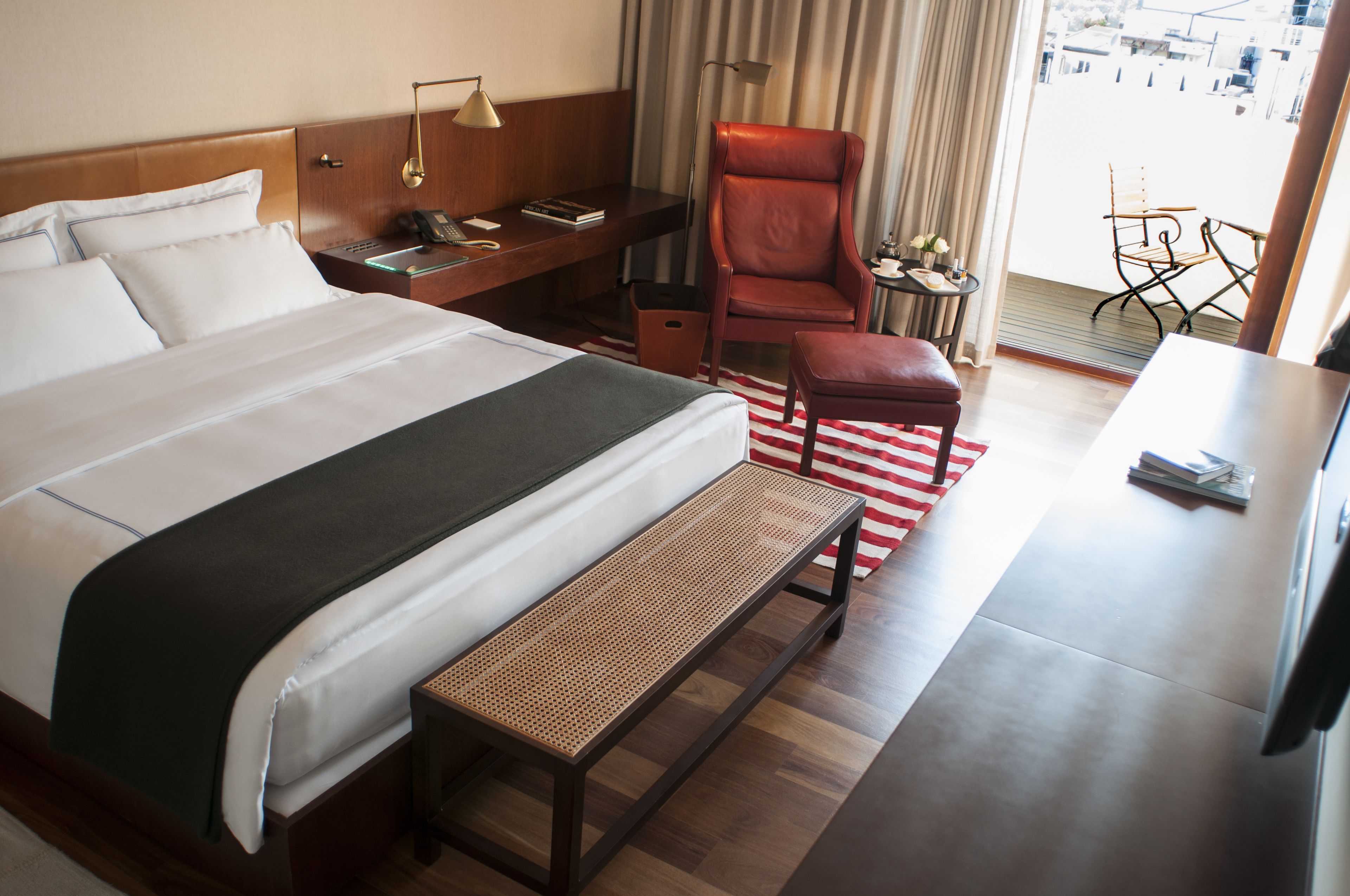 A hotel room with a large bed, a desk with a lamp, a red chair, a small table with a lamp, and a view of the city through the window.