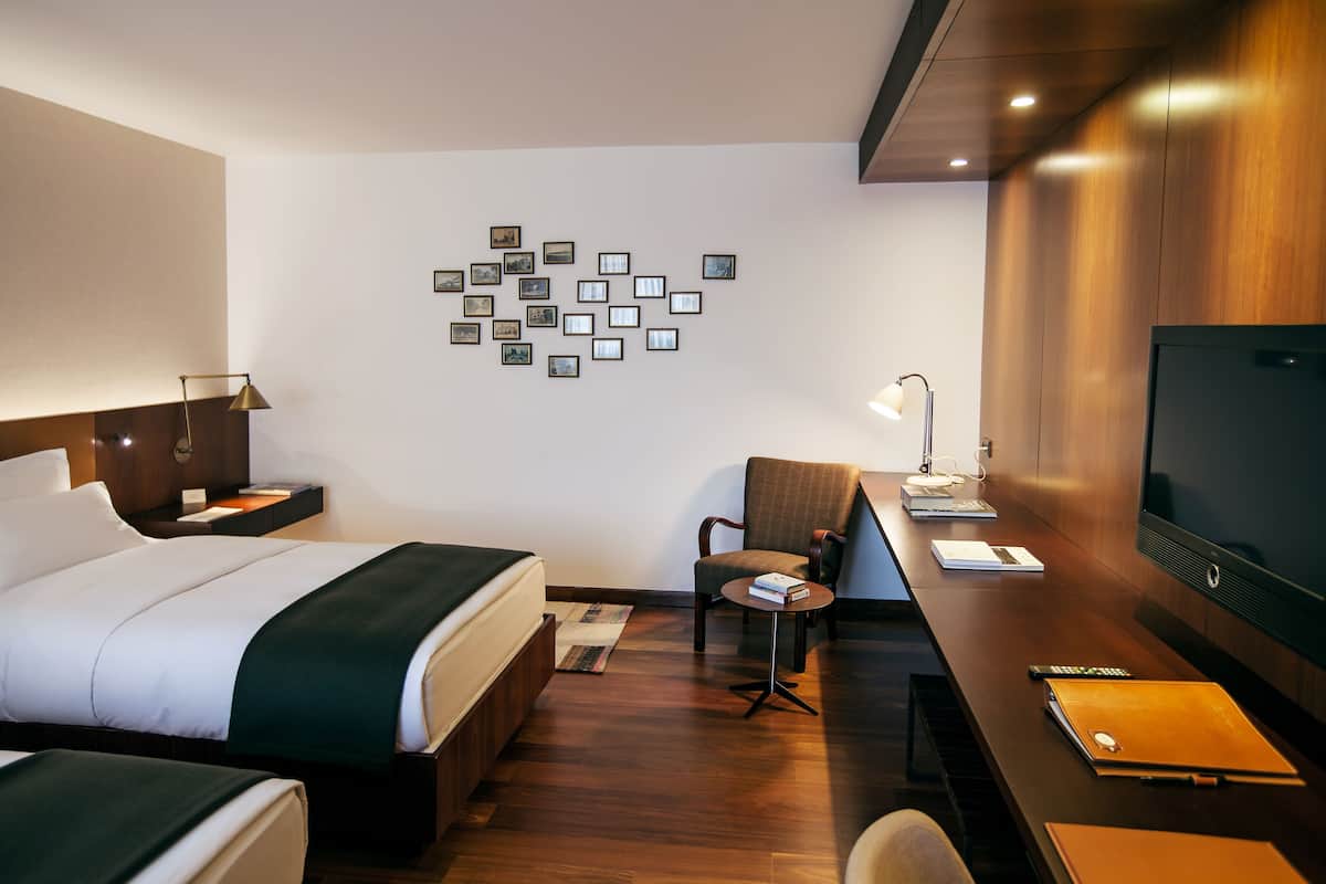 A hotel room with a bed, a desk with a chair, a TV, and a wall with framed pictures.