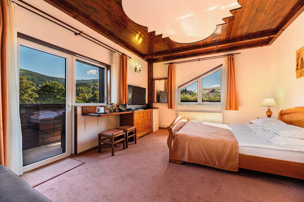 A bedroom with a bed, a desk, a chair, a television, and large windows.