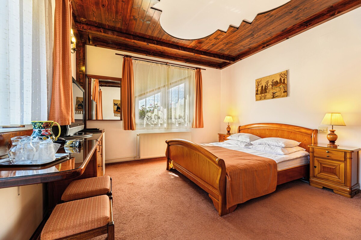A hotel room with a wooden bed, a desk with a chair, a window with curtains, a radiator, and a painting on the wall.