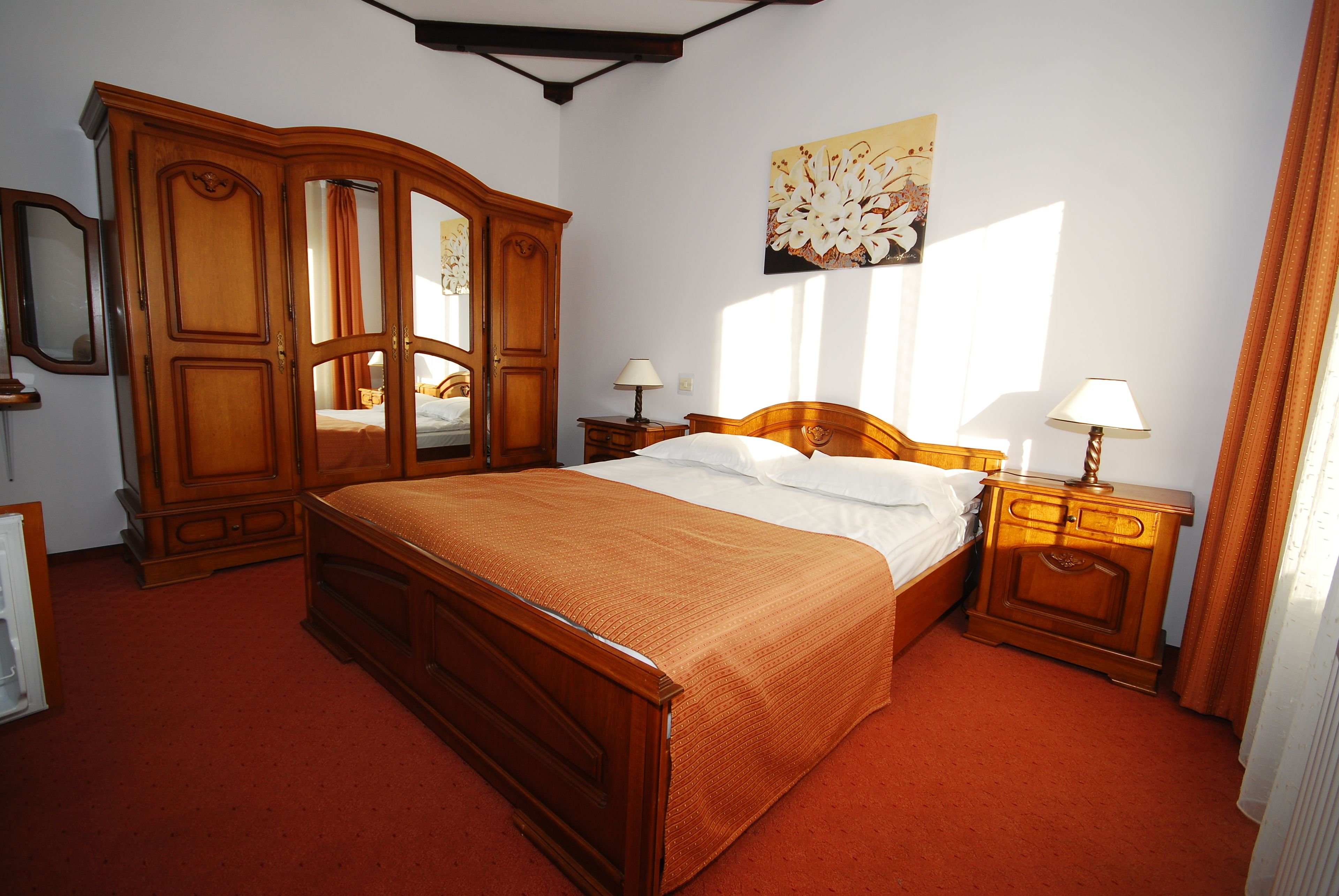 Un dormitorio con cama de madera, un armario, dos mesitas de noche con lámparas y un cuadro grande con motivos florales en la pared.