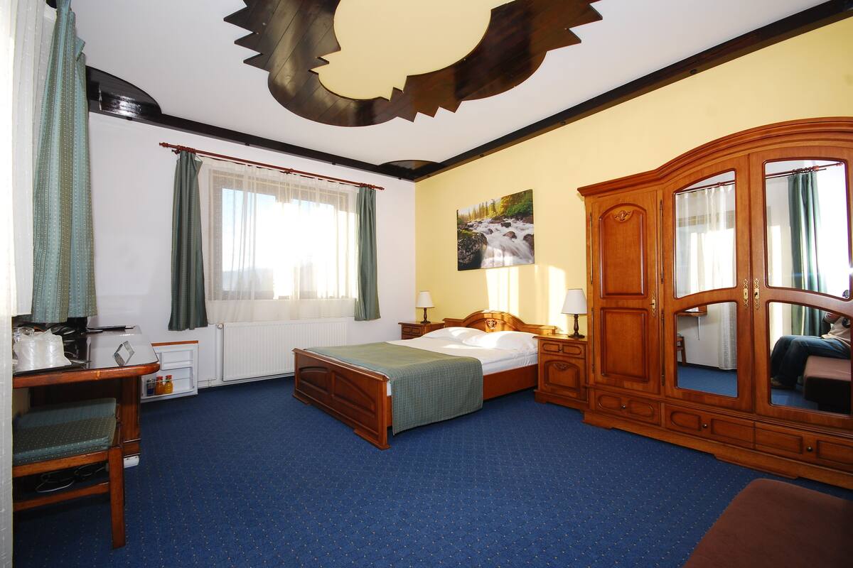 A hotel room with a bed, a wooden wardrobe, a desk with a chair, a window with curtains, and a painting on the wall.