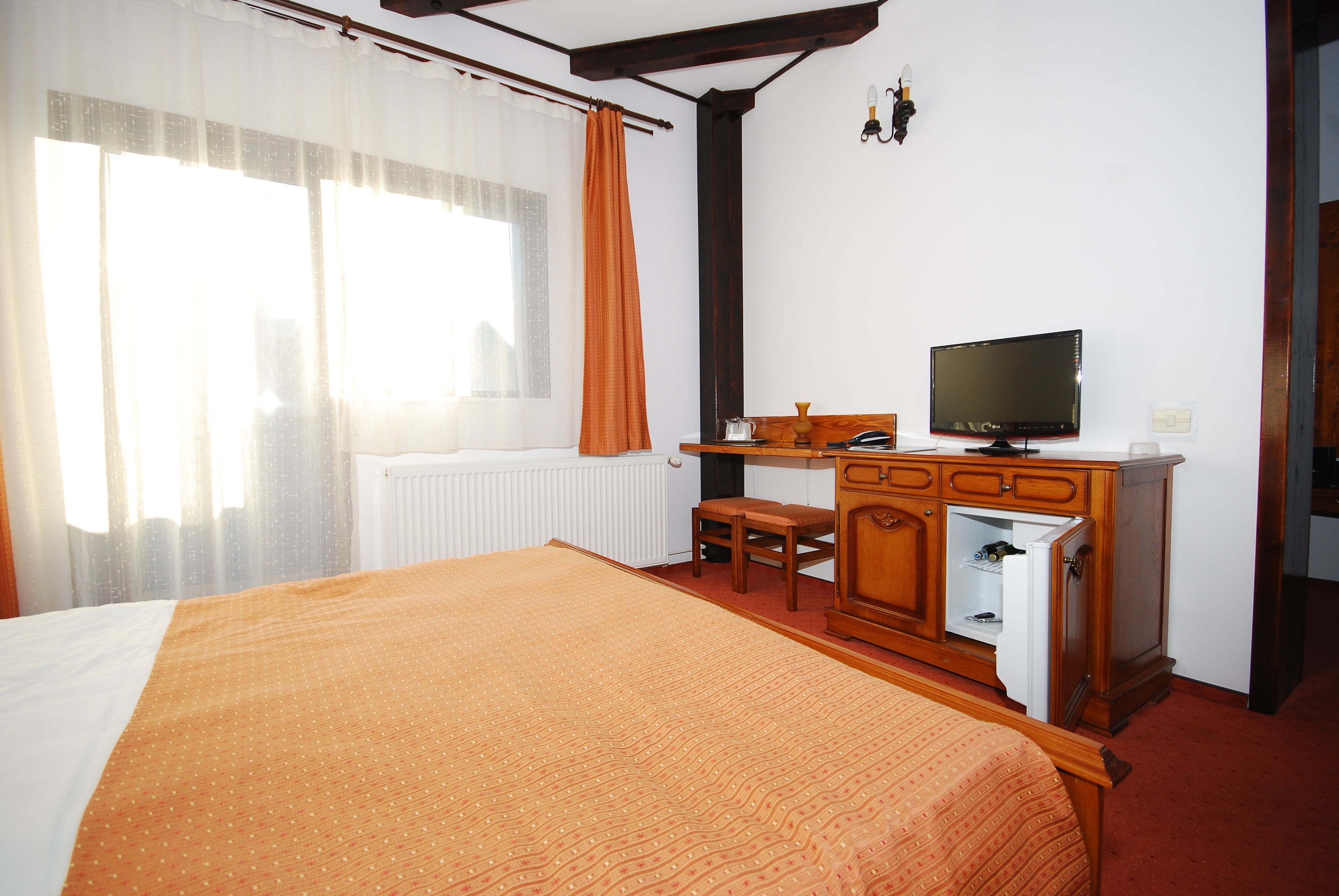 Quarto de hotel com cama, escrivaninha, televisão, radiador e janela com cortinas.
