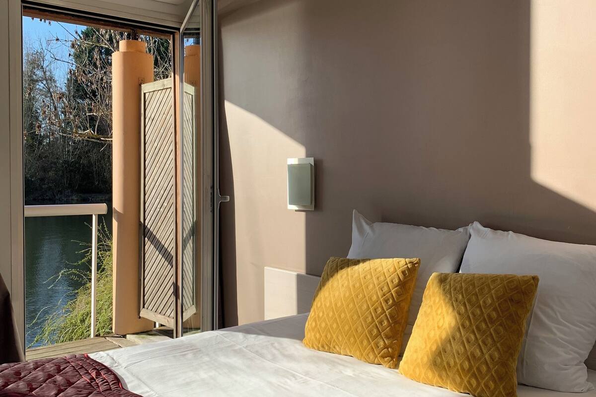 Habitación de hotel con cama, ventana con vistas a árboles y agua, unidad de aire acondicionado y una lámpara fijada a la pared.