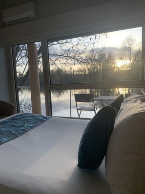 A bedroom with a bed, pillows, a view of a lake, and a chair outside the window. - Hôtel de l'Ile du Saussay (Itteville)