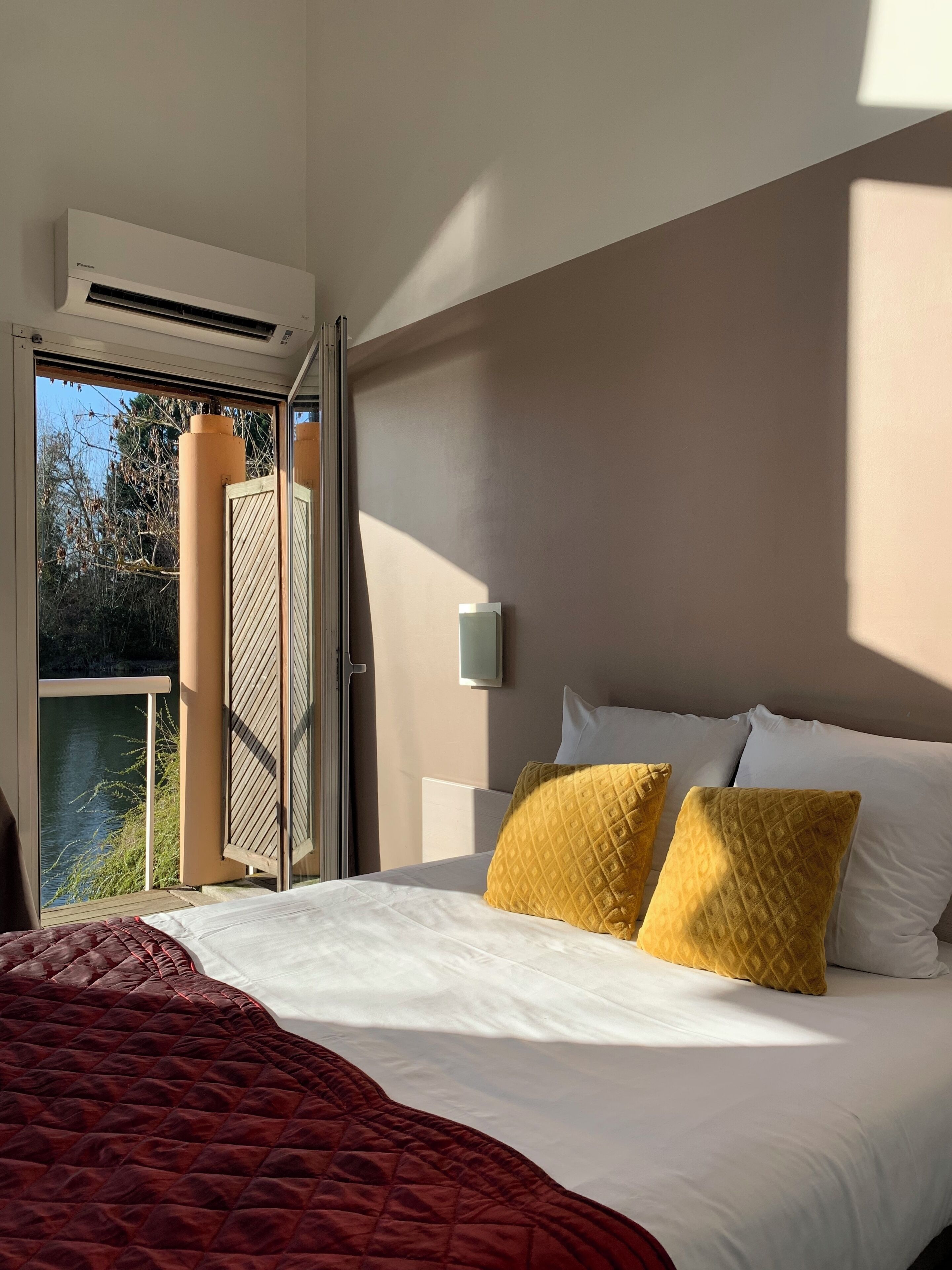 A hotel room with a bed, a window with a view of trees and water, an air conditioning unit, and a wall-mounted light.