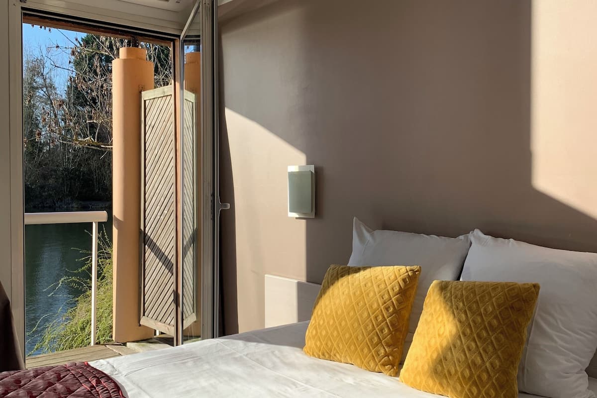 Habitación de hotel con cama, ventana con vistas a árboles y agua, unidad de aire acondicionado y una lámpara fijada a la pared.